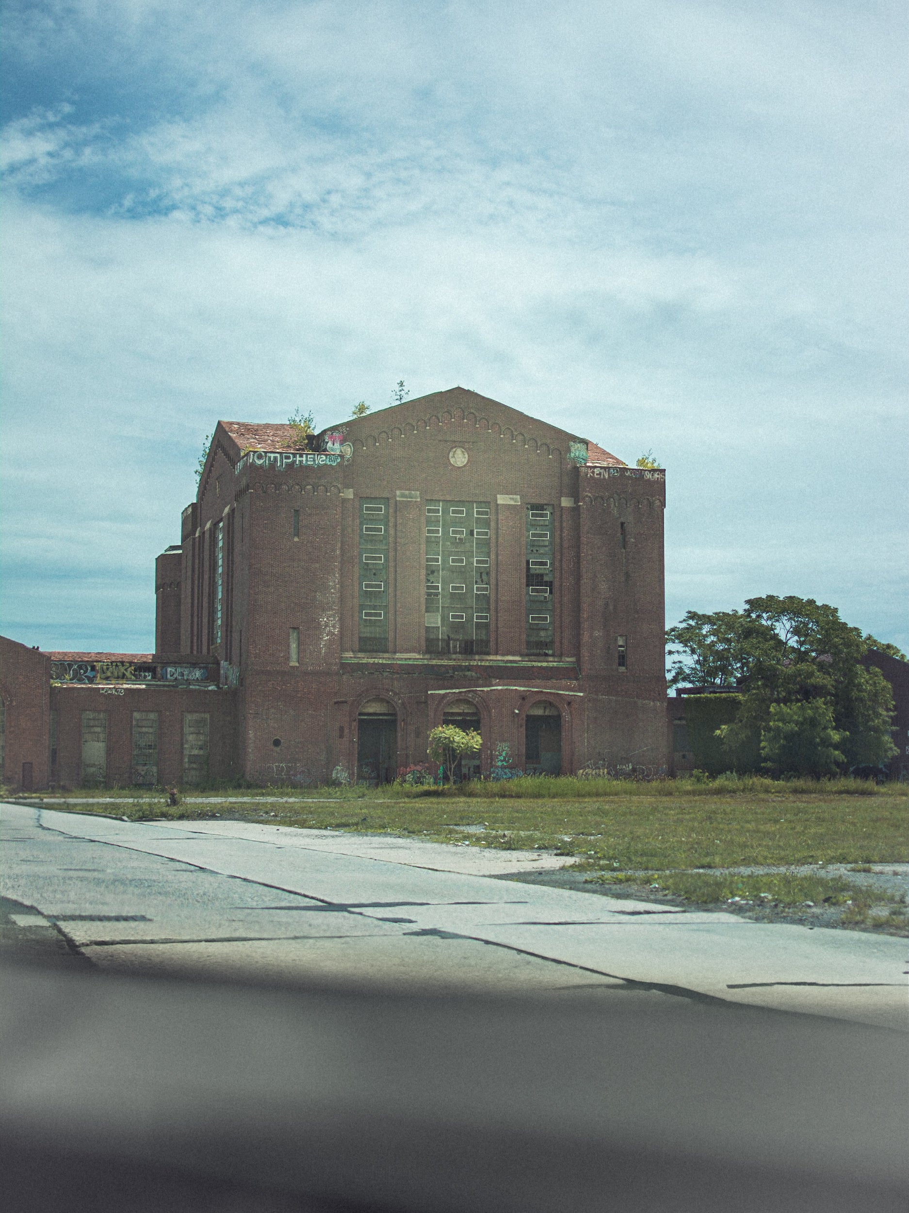 Abandoned brick building with overgrown grass and trees under a cloudy sky.